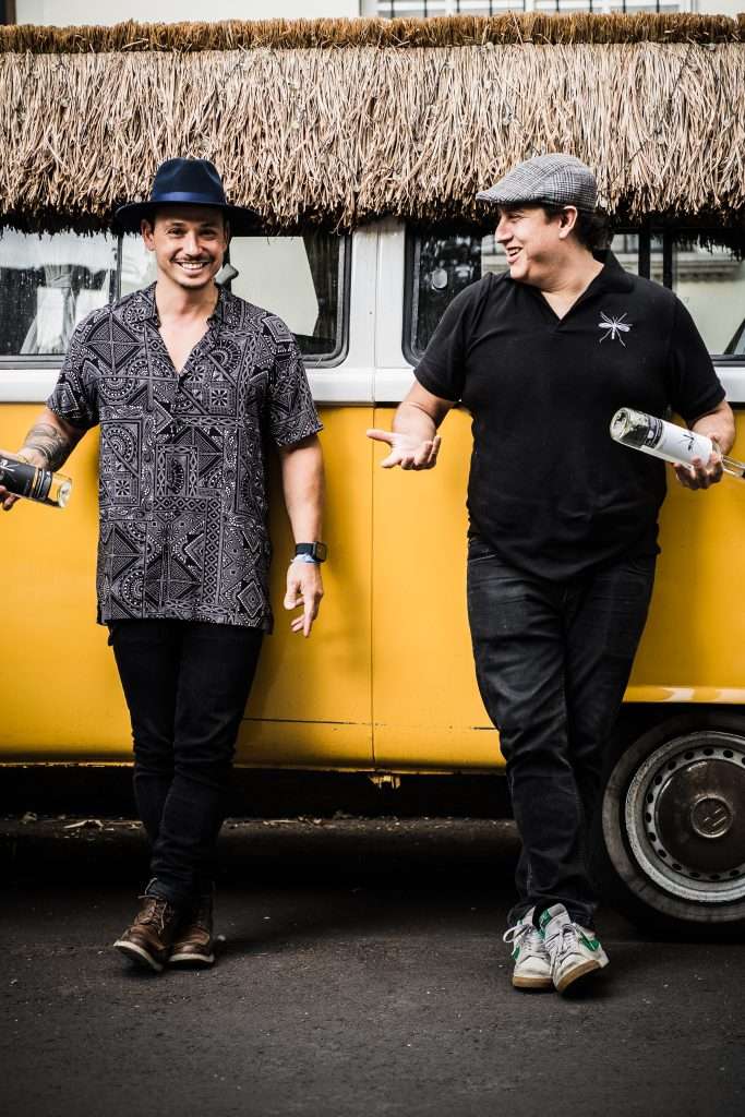 Zancudo Mezcal co-founders Andrés and Santos standing and smiling next to a classic yellow Volkswagen van outdoors.